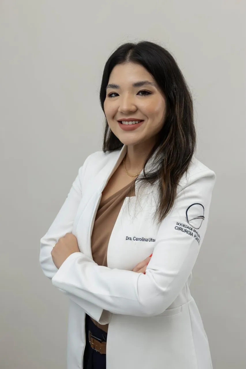 Dra. Carolina Utsunomiya de jaleco branco, sorrindo, com os braços cruzados